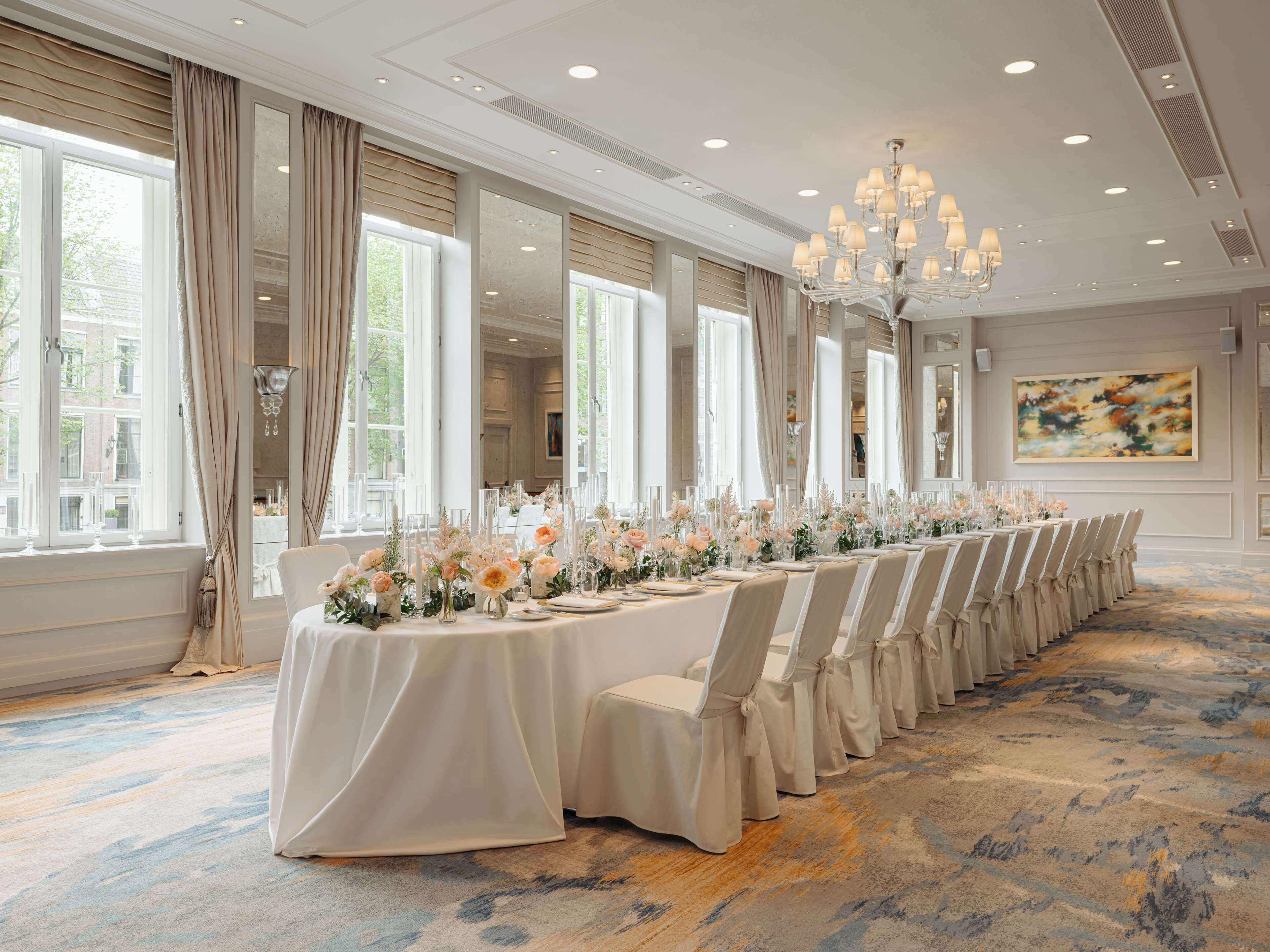 A long banquet table with chairs, set for a formal event, in a well-lit room with large windows and a chandelier.