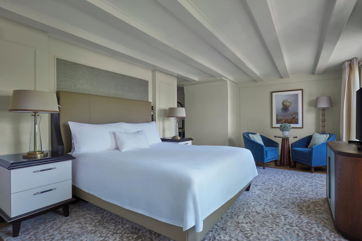 A hotel room with a large bed, two bedside tables with lamps, a small round table, two blue armchairs, and a painting on the wall.