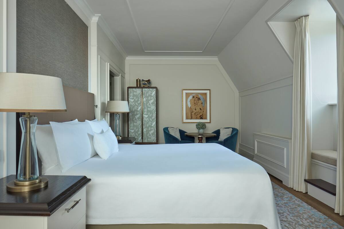 A hotel room with a large bed, bedside lamps, a small table with chairs, a decorative mirror, and a framed picture on the wall.