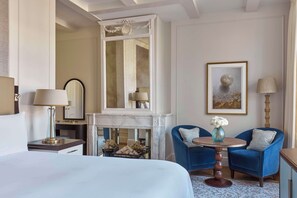 A hotel room with a large bed, a fireplace, two blue armchairs, a round table with a vase, and a framed picture on the wall. - Waldorf Astoria Amsterdam (Amsterdam)