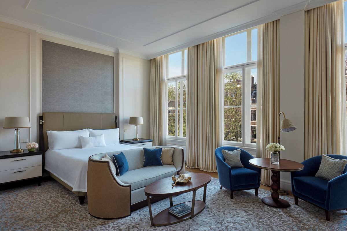 A hotel room with a large bed, two armchairs, a coffee table, and a small table with a vase of flowers.