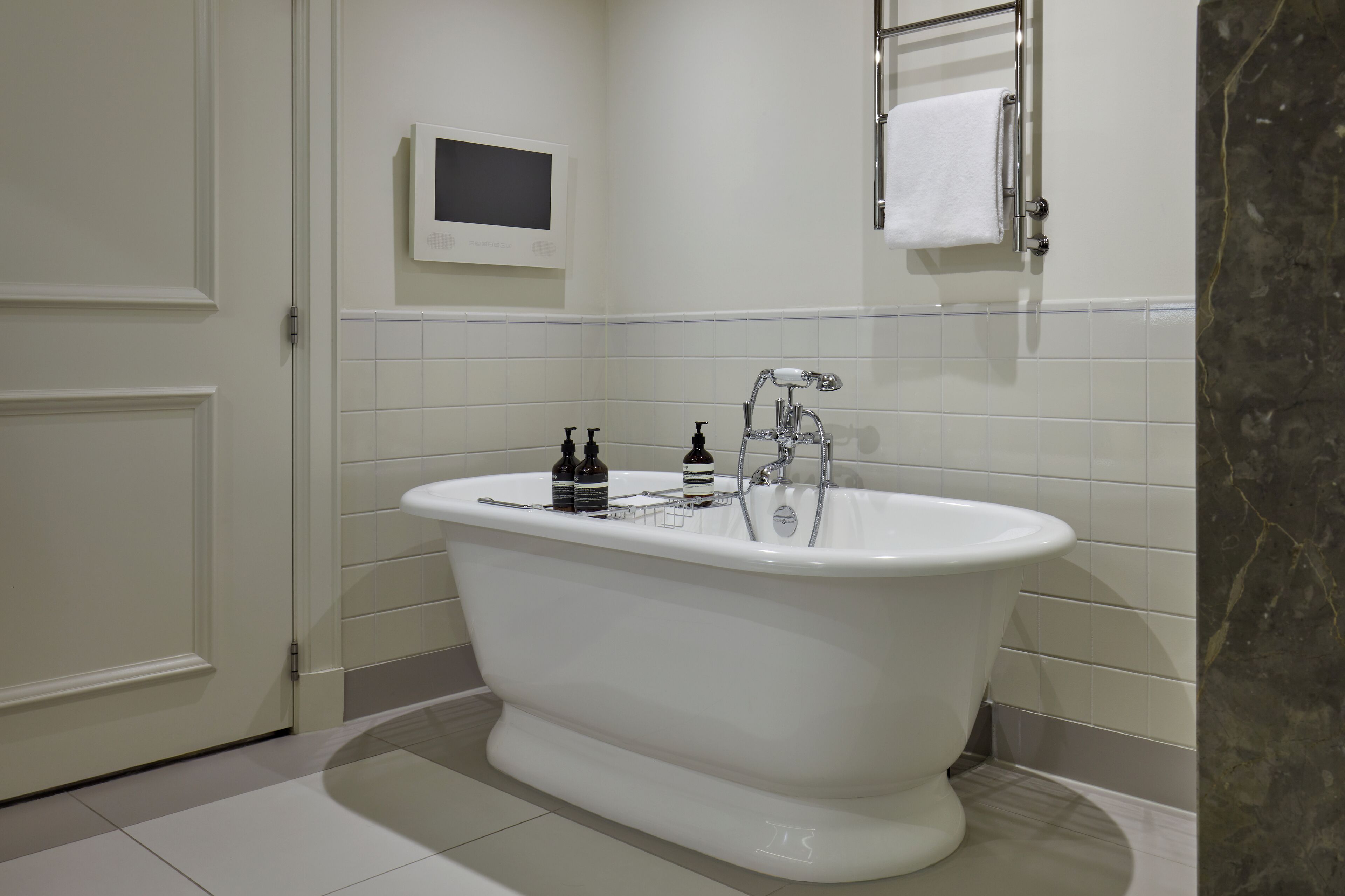A modern bathroom with a freestanding bathtub, white tiles, a mounted TV, and a towel rack.
