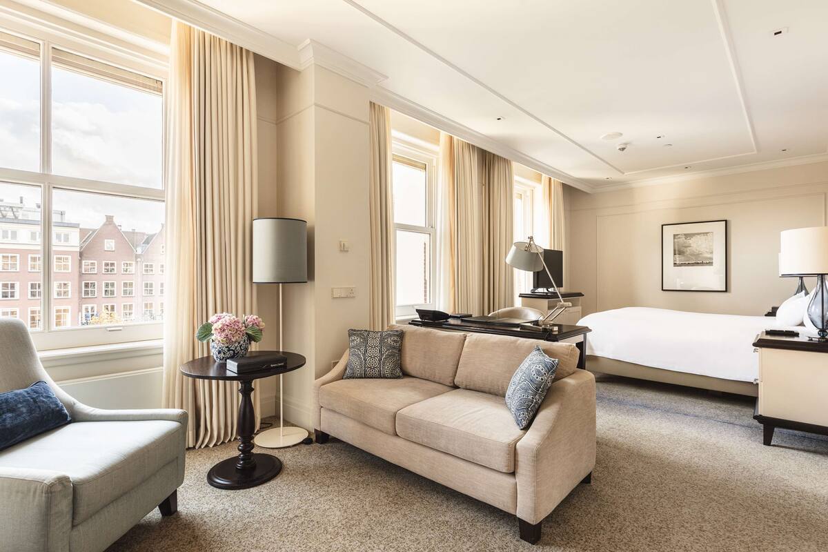 A hotel room with a bed, a sofa, a desk, and a view of buildings outside the window.