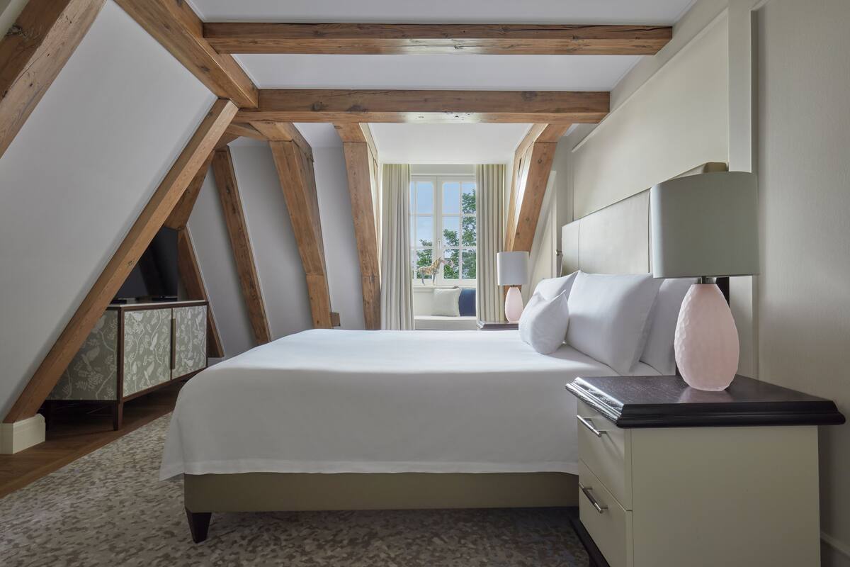 A bedroom with a large bed, a nightstand, a lamp, and a view of the outdoors through a window.