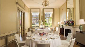 A dining room with a round table set for a meal, large windows with a view of buildings, and a chandelier.