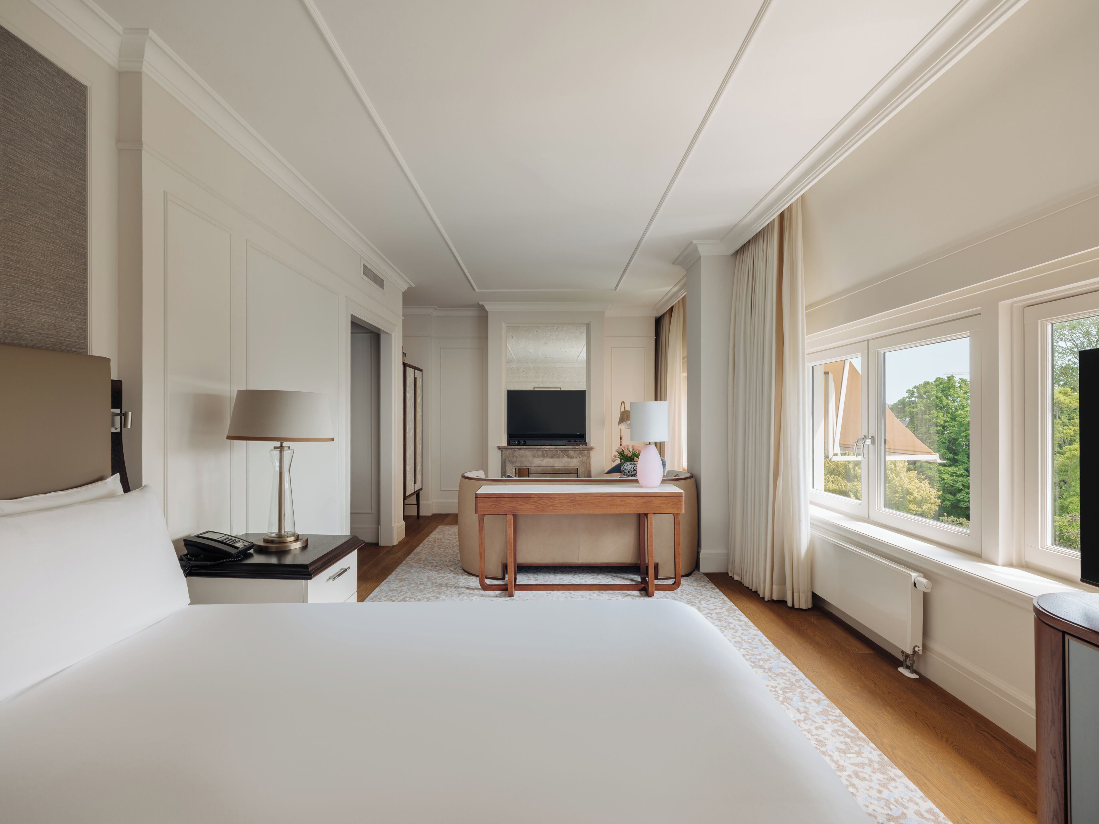 A hotel room with a large bed, a desk with a TV, and a window with a view of greenery.