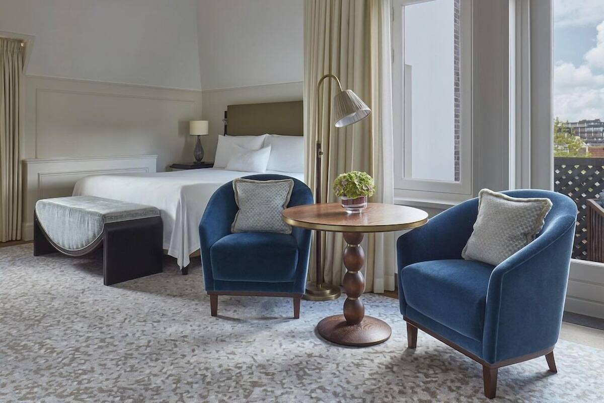 A hotel room with a bed, two blue armchairs, a small round table with a plant, a lamp, and a view of the city.