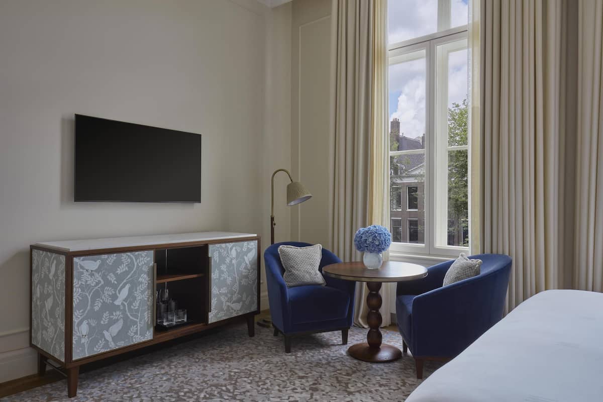 A hotel room with a bed, a TV mounted on the wall, a small table with chairs, a cabinet with patterned doors, and a window with curtains.