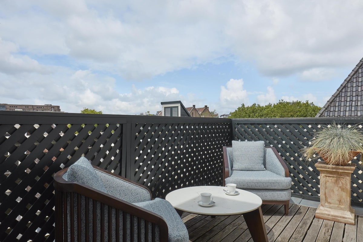 An outdoor seating area with a wooden deck, two grey upholstered chairs, a small round table, and a potted plant.
