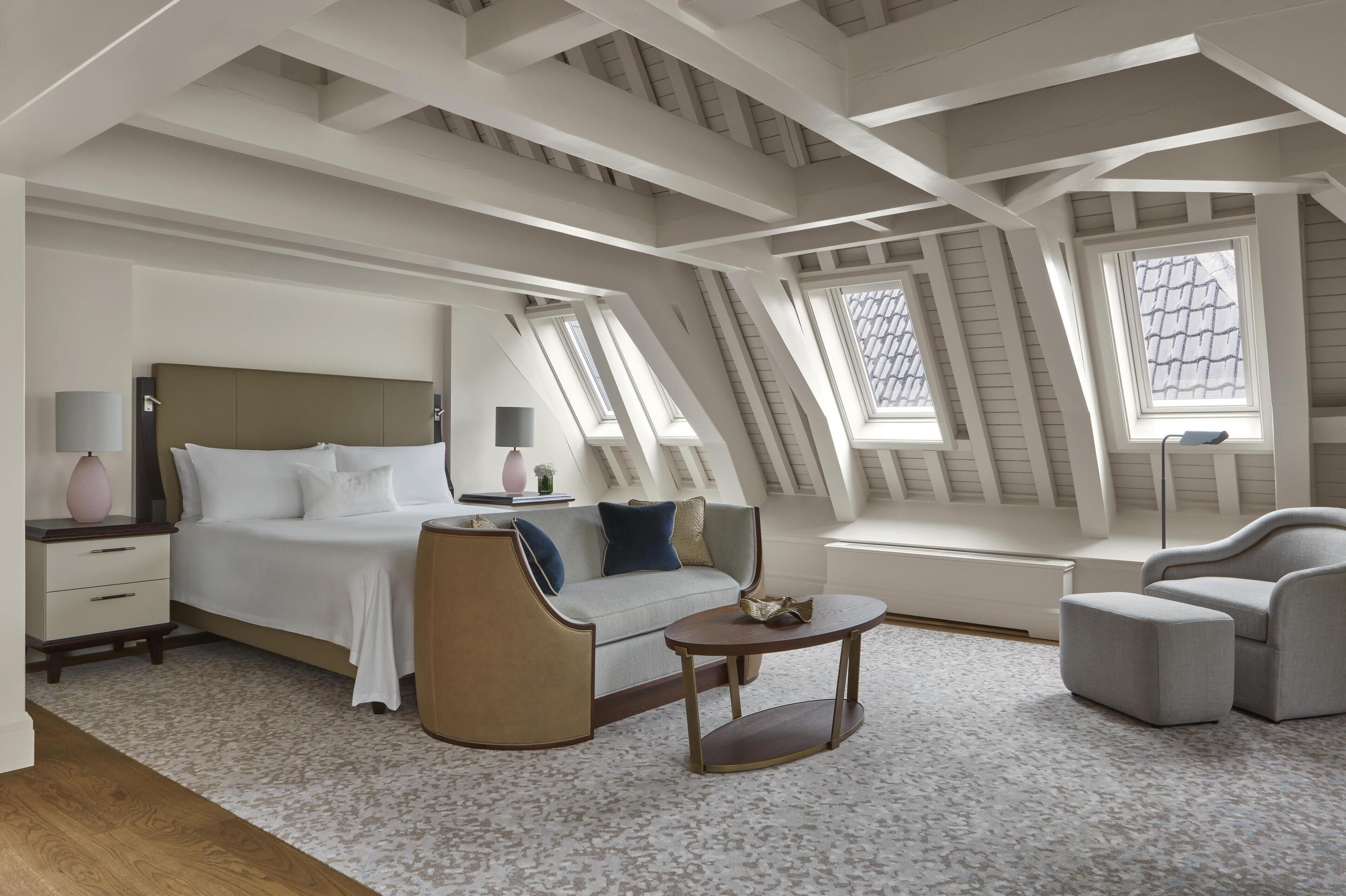 Una camera da letto moderna con un letto, un divano, una poltrona e un tavolino. La stanza ha travi a vista con lucernari e un tappeto decorato.
