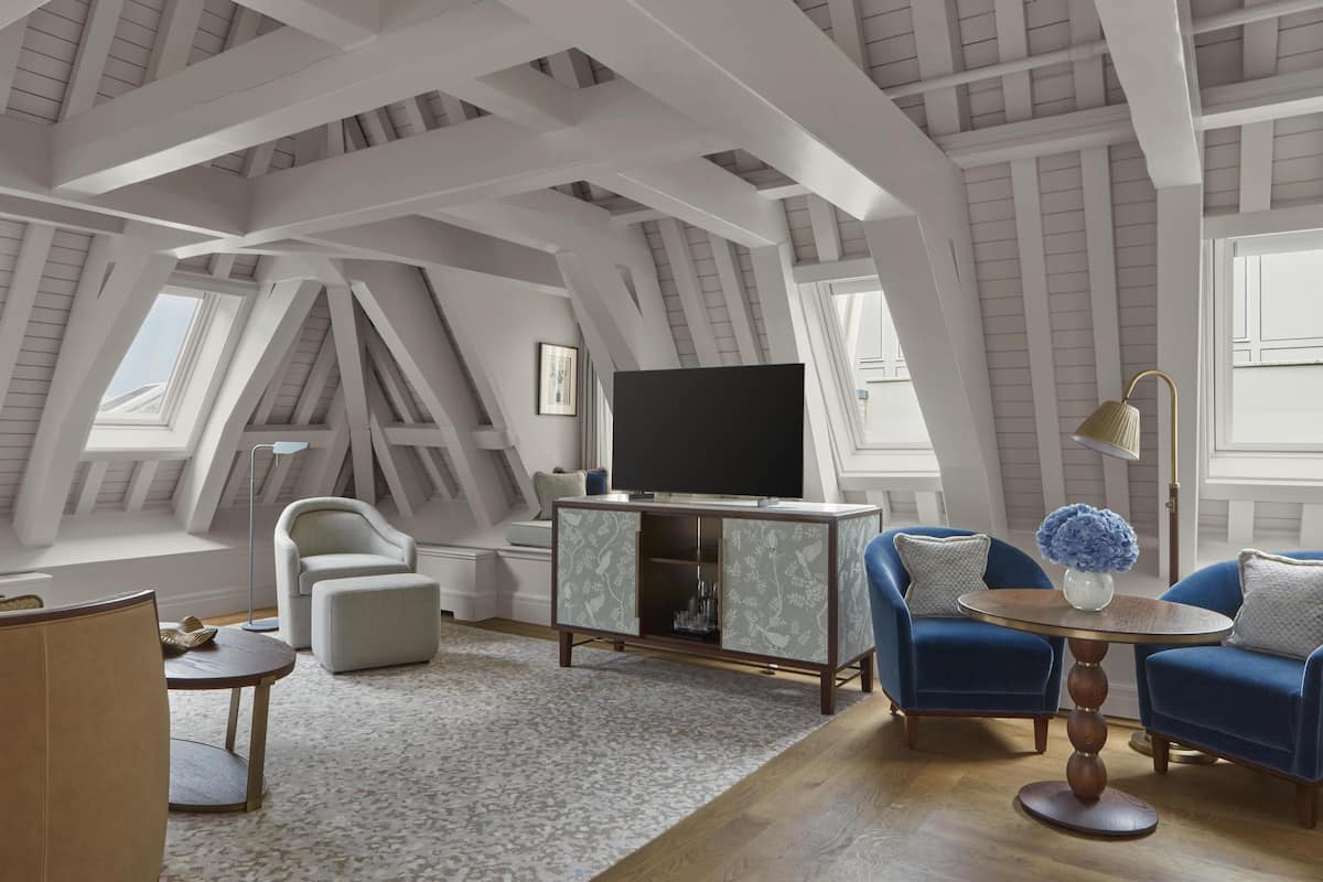 A modern loft bedroom with a sloped ceiling, built-in TV, and a cozy seating area with a round coffee table and blue armchairs.