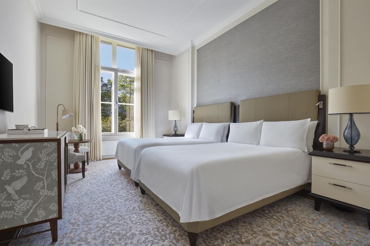 A hotel room with two beds, a desk, a chair, a TV, and a window with curtains.