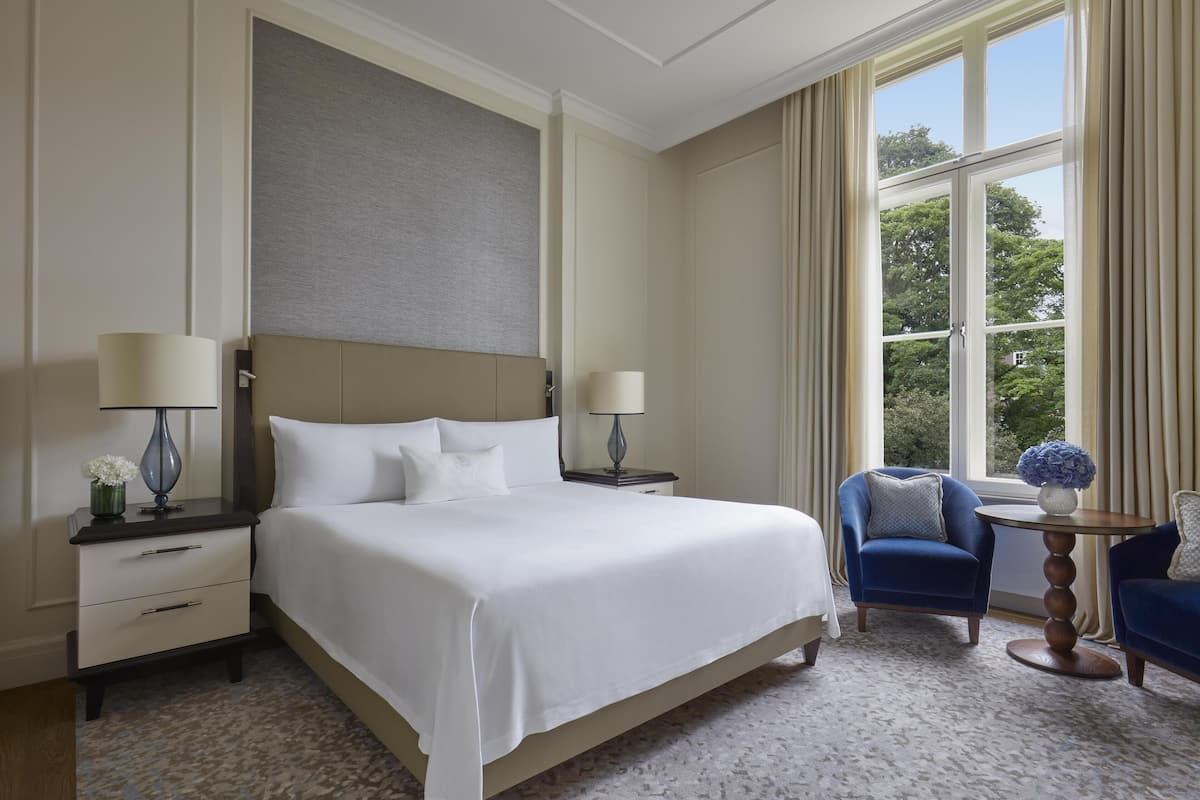 A hotel room with a large bed, two bedside tables with lamps, a small round table with a vase, two blue armchairs, and a window with a view of trees.