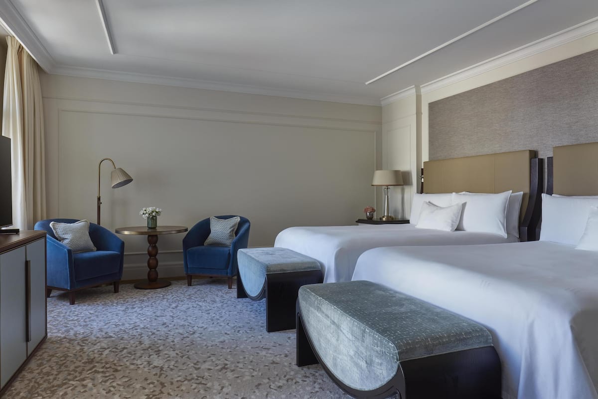 A hotel room with two beds, a desk, two chairs, a small table, and a lamp.