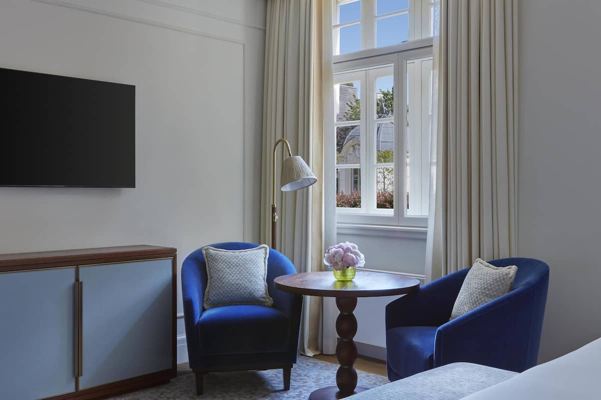A room with a large flat-screen TV mounted on the wall, two blue armchairs, a small round table with a vase of flowers, and a window with curtains.