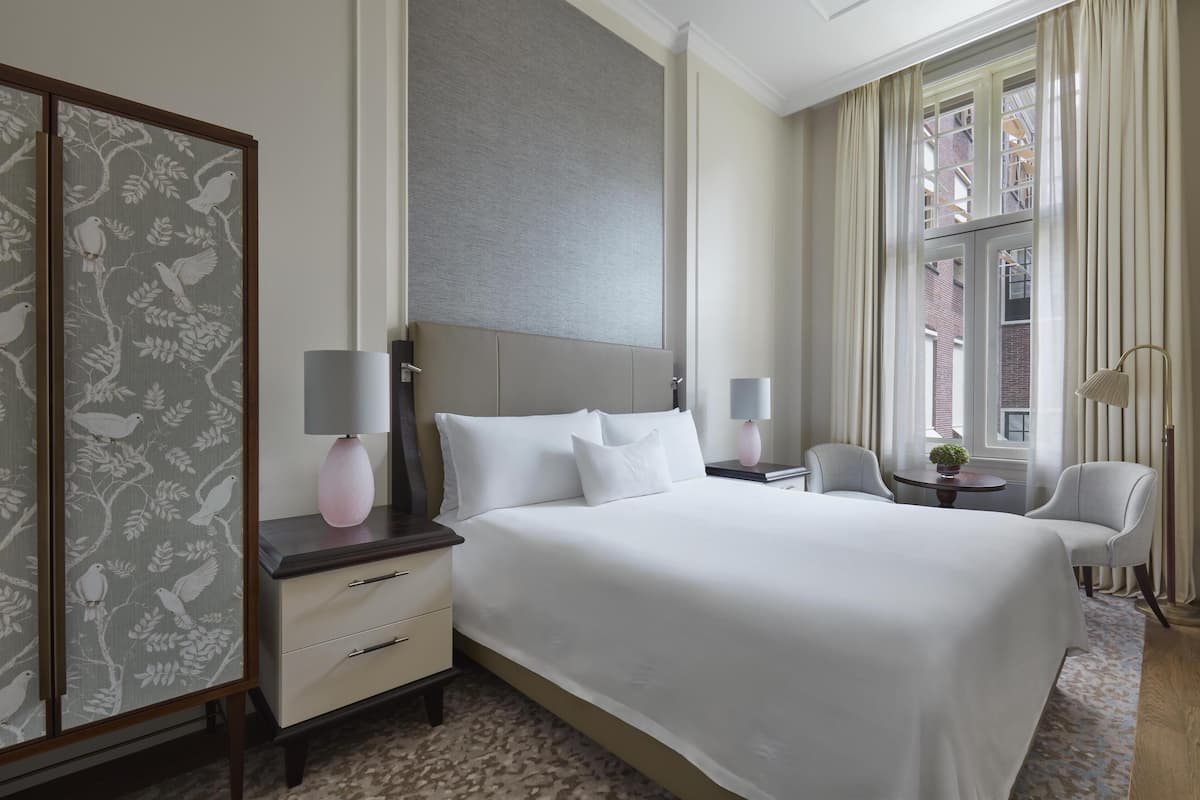 A hotel room with a large bed, two bedside tables with lamps, a chair, a small table with a plant, and a view of the city through the window.