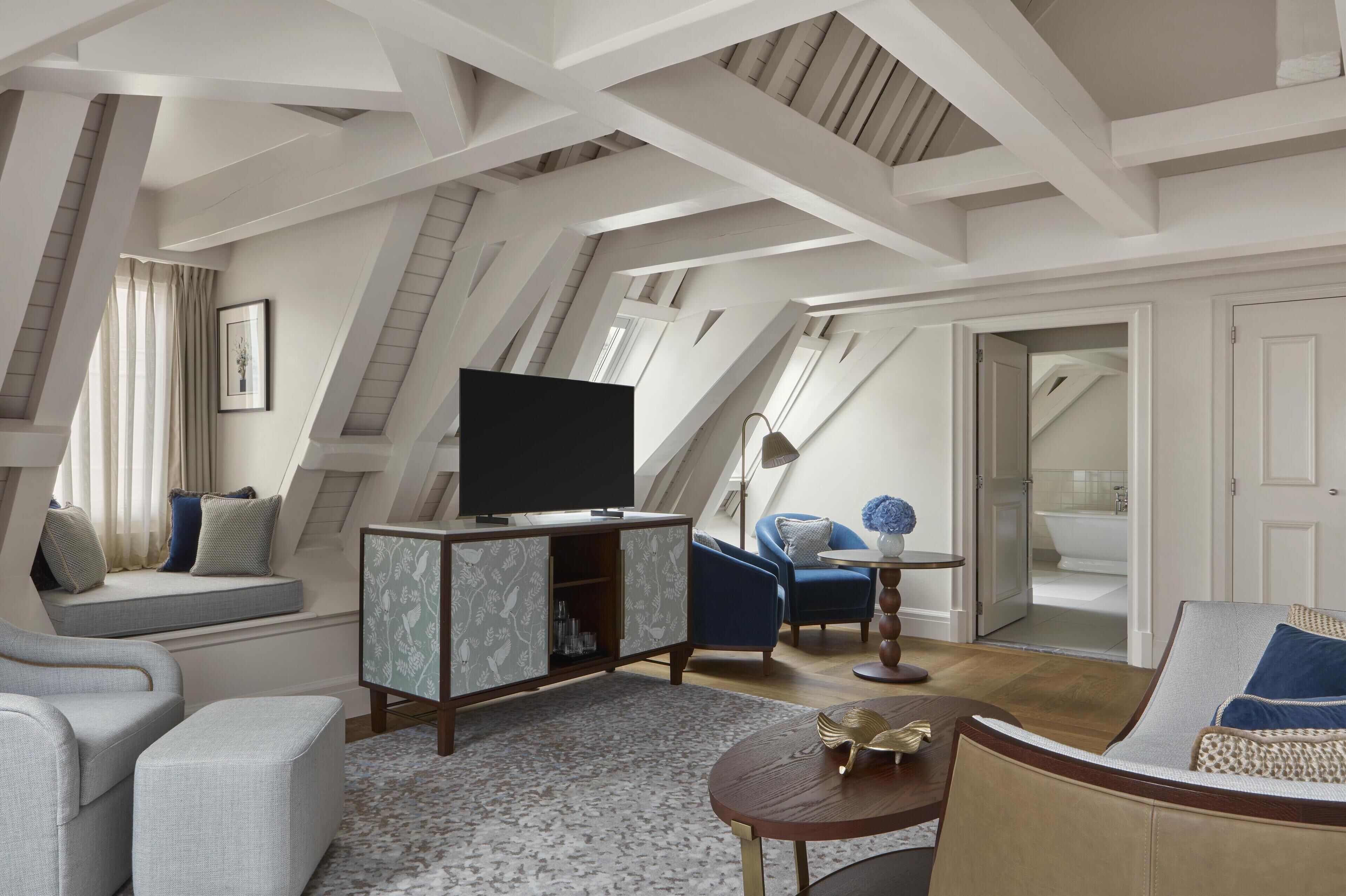 A modern loft bedroom with a sloped ceiling, built-in seating, a flat-screen TV, and a bathroom visible through an open door.