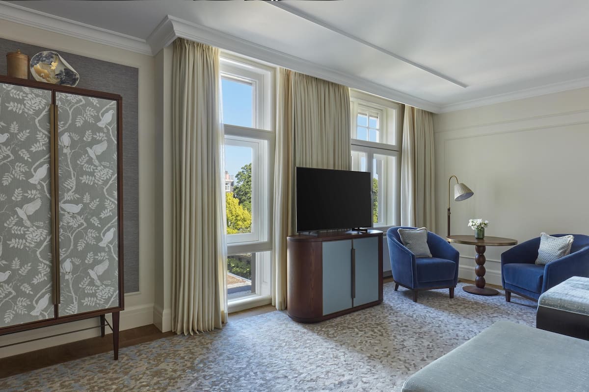 A hotel room with a large window, a flat-screen TV, two blue armchairs, a round side table, and a cabinet with a floral pattern.