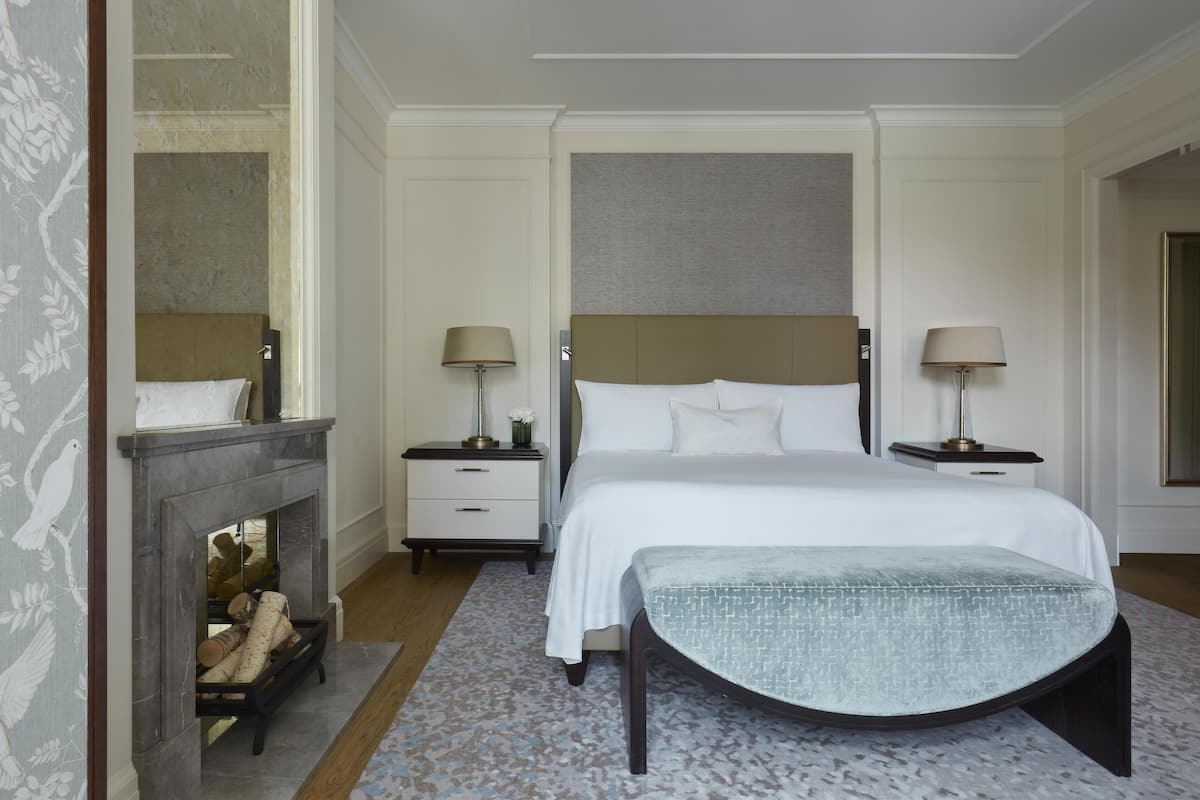A bedroom with a large bed, two bedside tables with lamps, a fireplace, and a carpeted floor.
