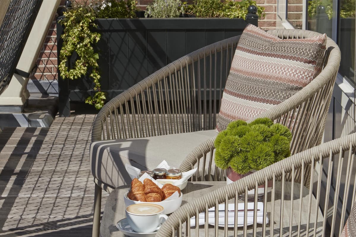 A wicker chair with a cushion, a small potted plant, a basket of croissants, and a cup of coffee on a balcony.