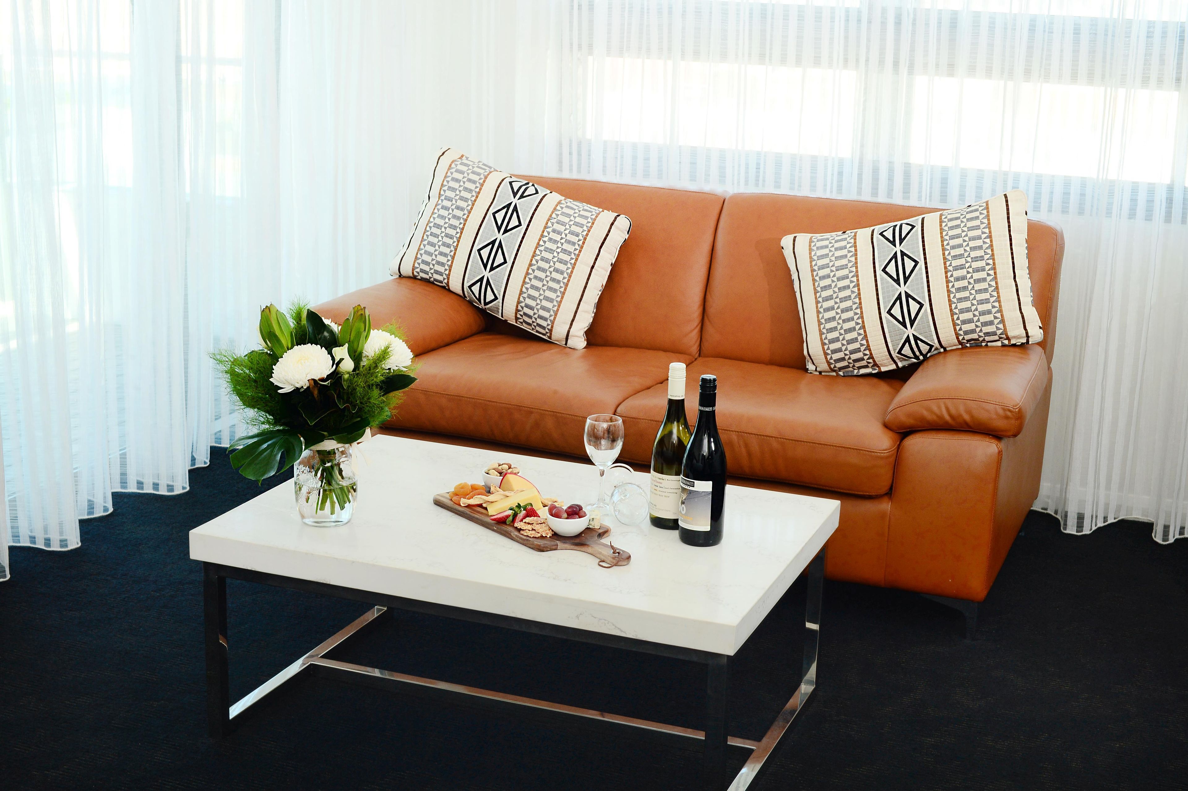 Una sala de estar con un sofá de cuero marrón, dos almohadones estampados, una mesa de centro blanca con un arreglo floral, una botella de vino y una copa.