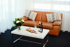 A living room with a brown leather sofa, two patterned pillows, a white coffee table with a floral arrangement, a bottle of wine, and a glass.