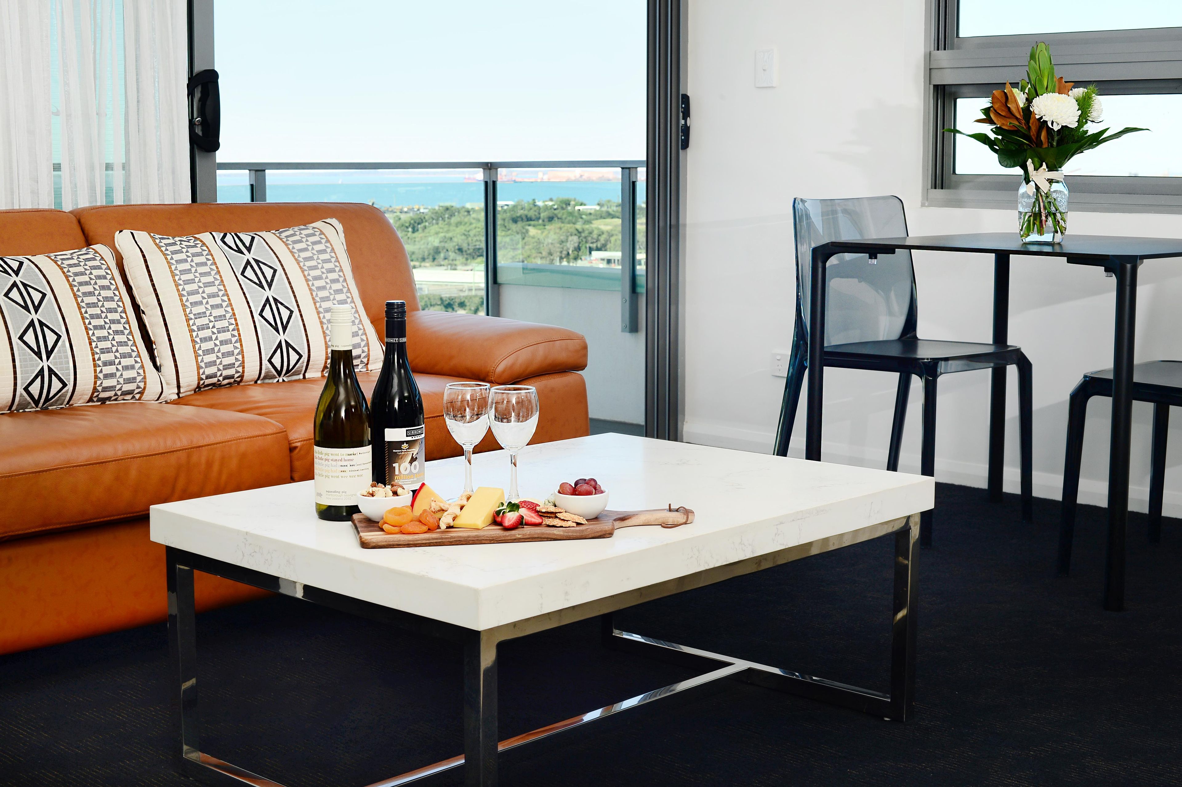 A modern living room with a leather sofa, a white coffee table with a bottle of wine, glasses, and a fruit platter.