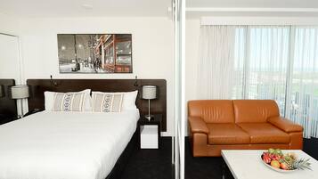 A hotel room with a large bed, a brown leather sofa, a small table with fruit, and a painting on the wall.