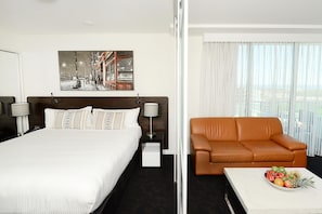 A hotel room with a large bed, a brown leather sofa, a small table with fruit, and a painting on the wall.