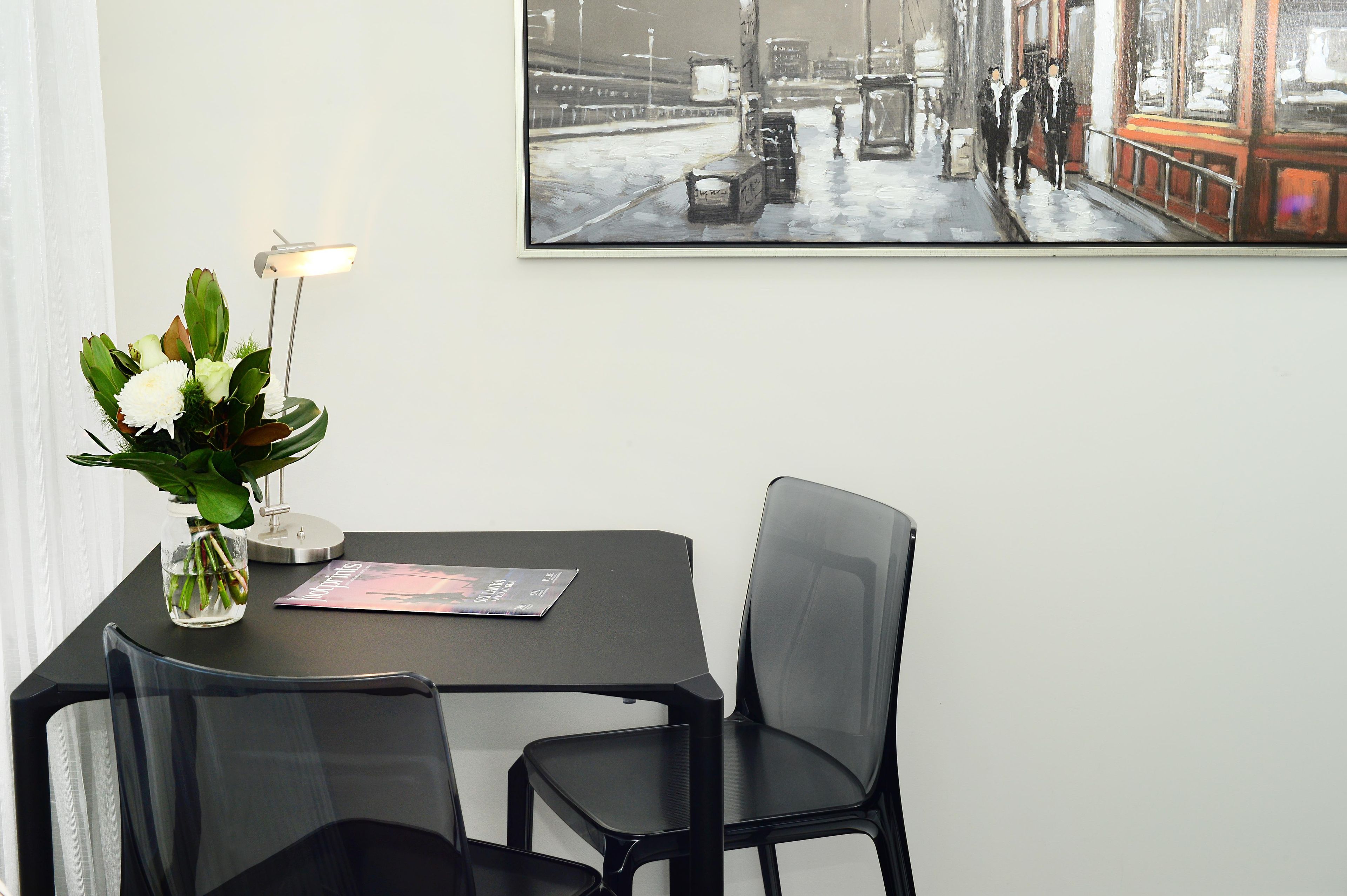 A table with a vase of flowers, a lamp, and a magazine, with a painting of a street scene on the wall.