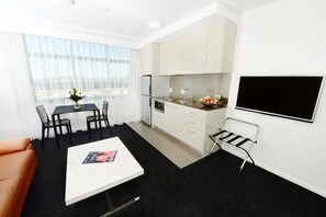 Un espacio habitable compacto con kitchenette, un comedor con mesa y sillas, un sofá negro y un televisor montado en la pared.