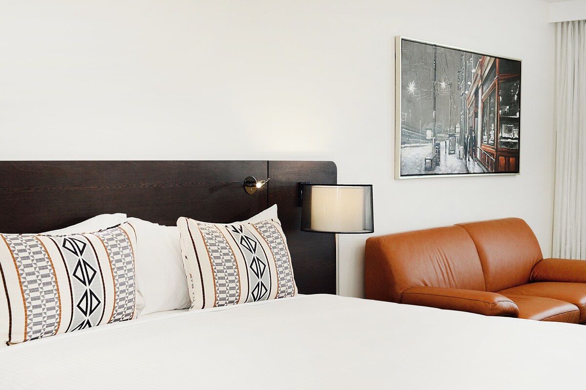 Una habitación de hotel con una cama, un sillón de cuero marrón, una mesita de noche con lámpara y un cuadro enmarcado en la pared.