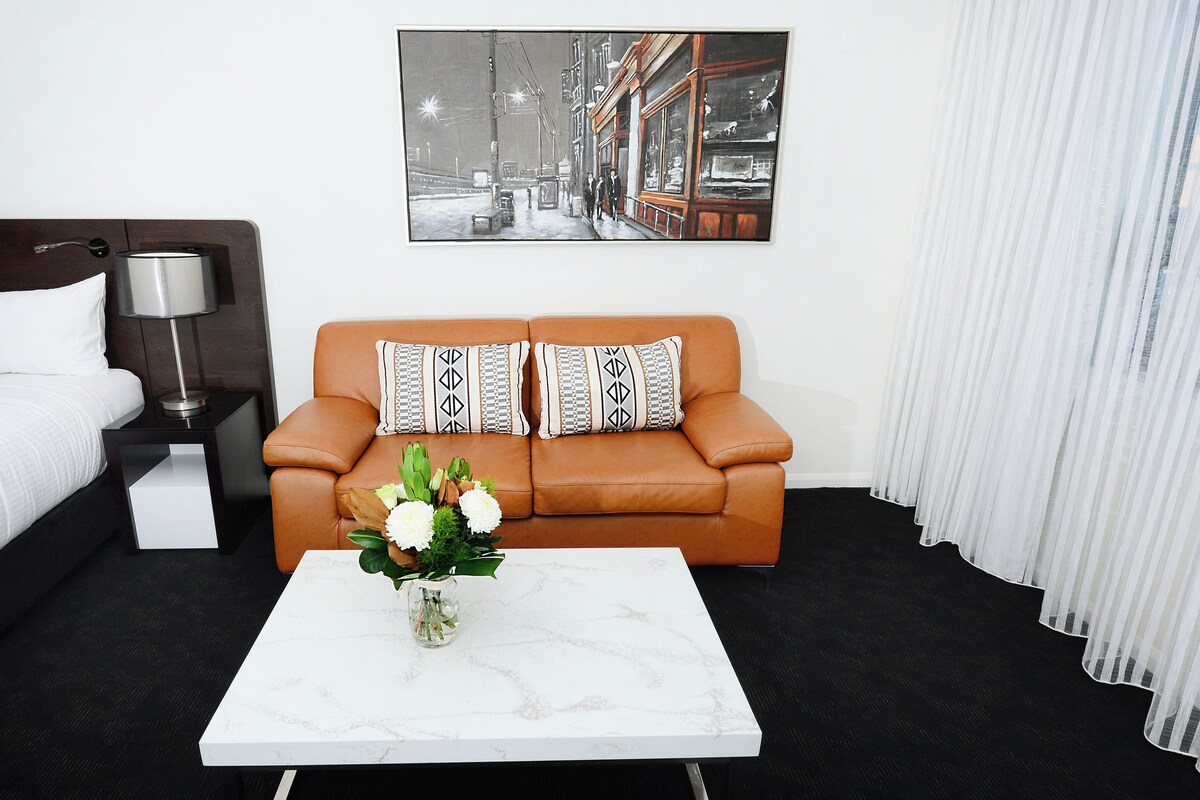Una habitación de hotel moderna con un sofá de cuero marrón, una mesa de centro de mármol blanco con un jarrón de flores, un cuadro grande con una vista urbana enmarcada y una mesita de noche negra con una lámpara.
