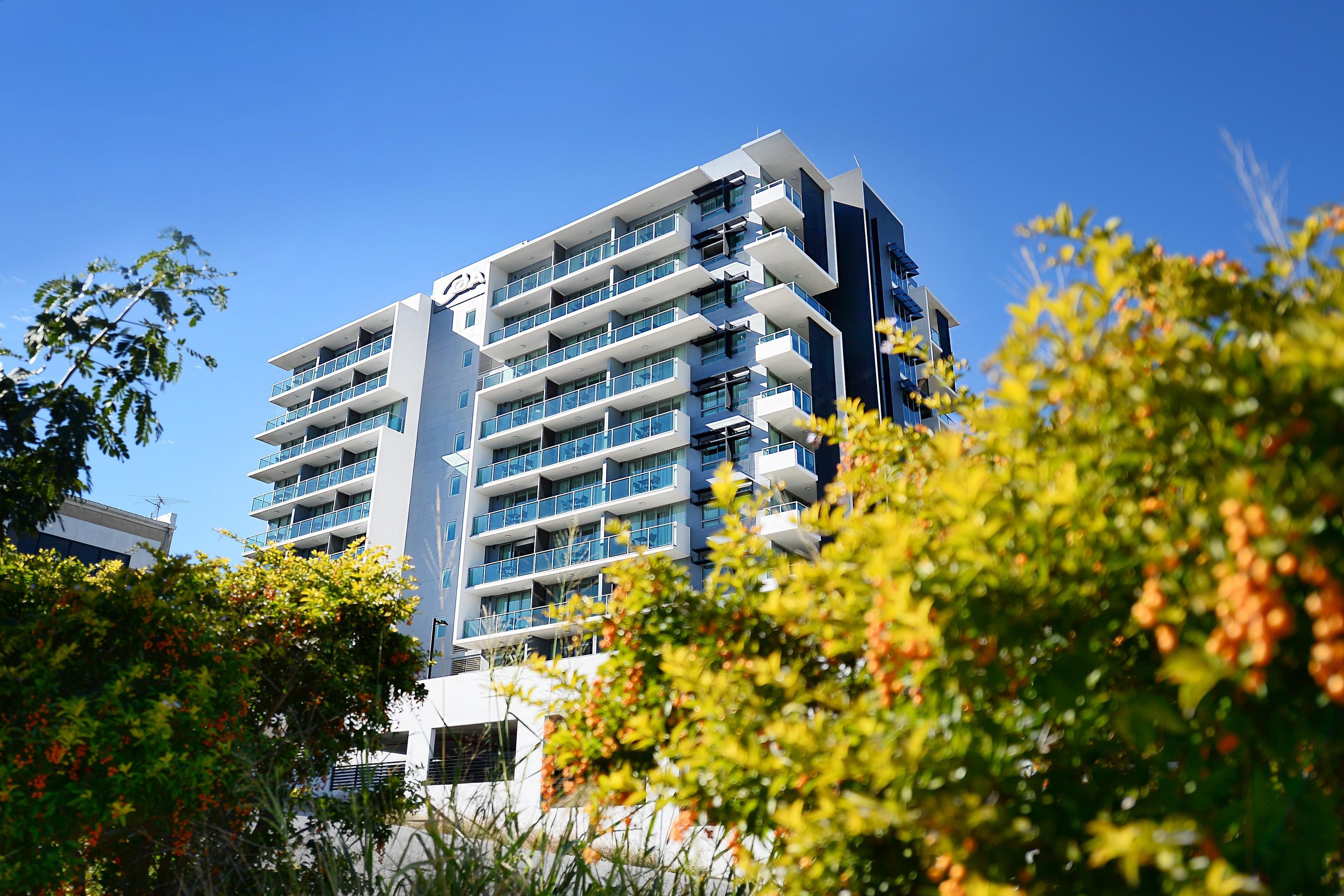 A multi-story building with balconies and large windows, surrounded by greenery.