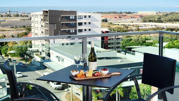 Un balcón con una mesa dispuesta para una comida, una botella de vino y vistas a la ciudad y al mar.