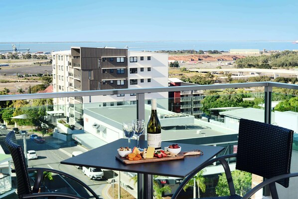 Un balcón con una mesa dispuesta para una comida, una botella de vino y vistas a la ciudad y al mar.