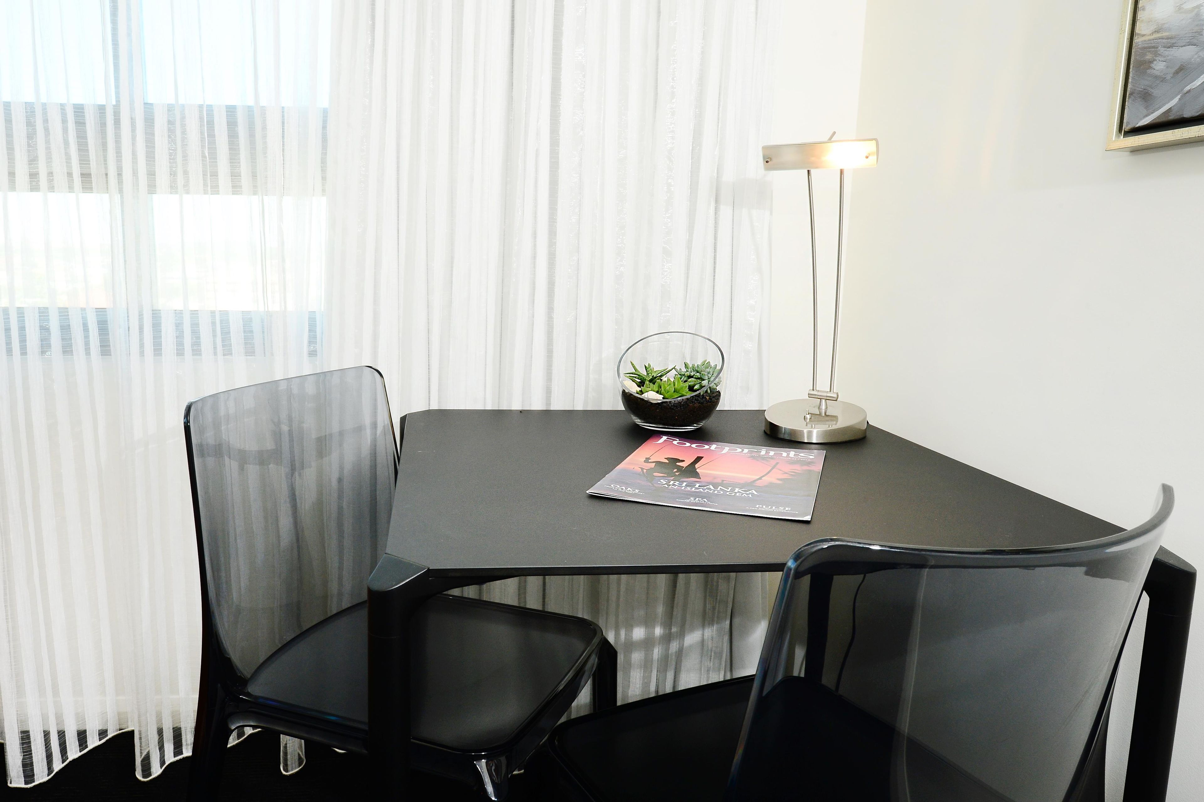 A modern office space with a glass table, two black chairs, a small potted plant, a lamp, and a magazine on the table.