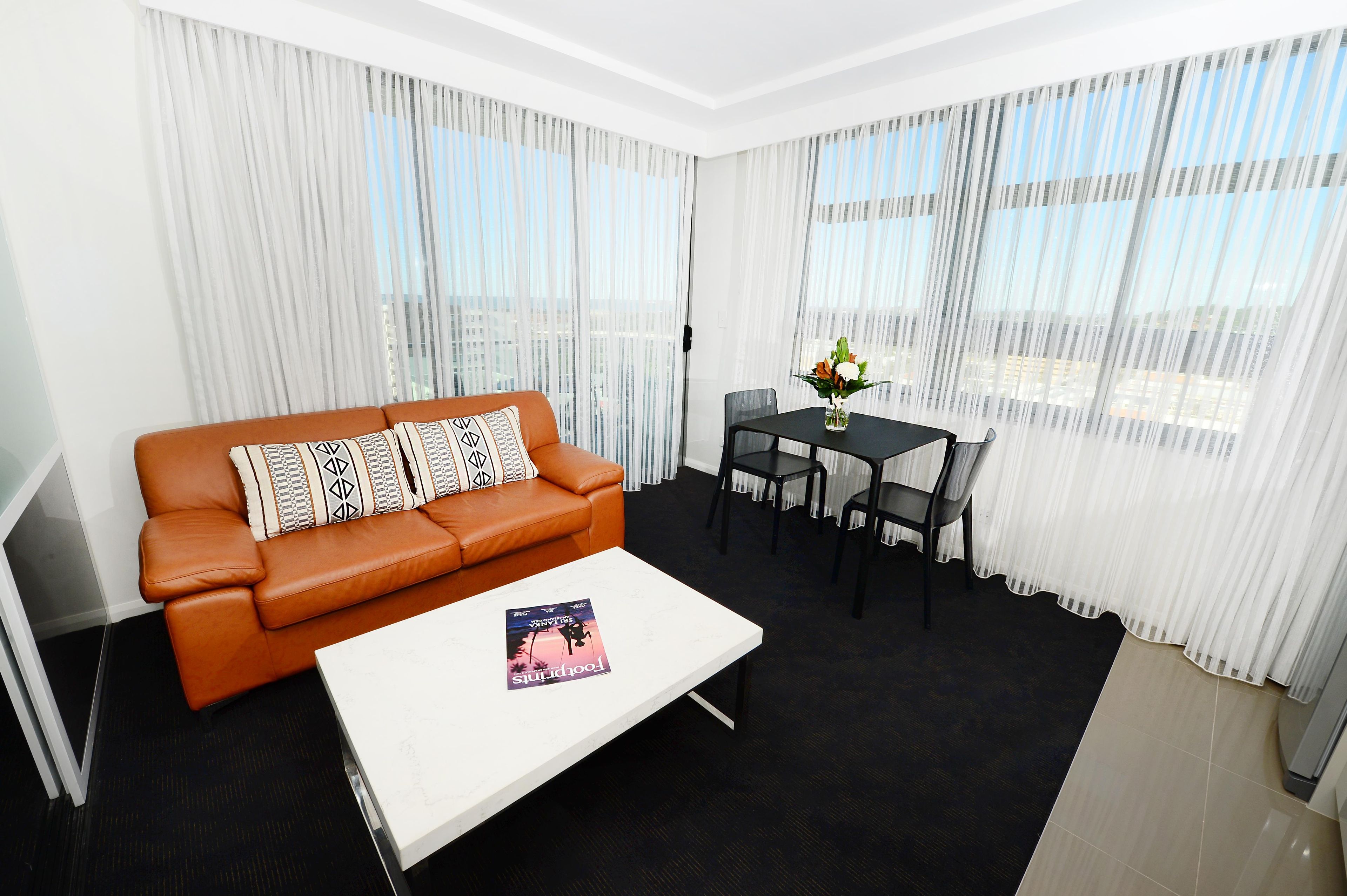 A modern living room with a brown leather sofa, a white coffee table with a book, a dining table with chairs, and sheer white curtains.