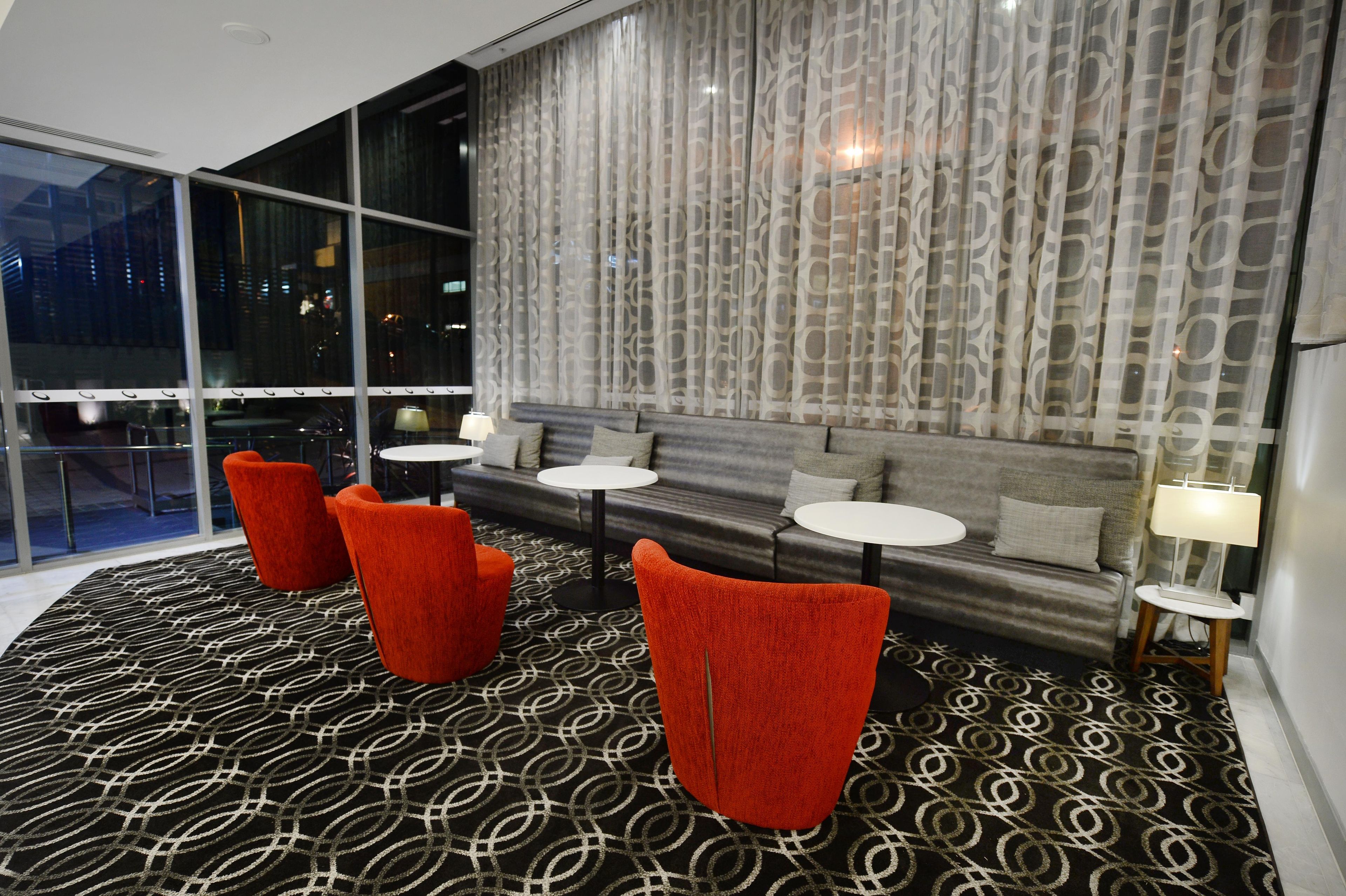 A modern hotel lobby with a large window, a patterned carpet, and red chairs.