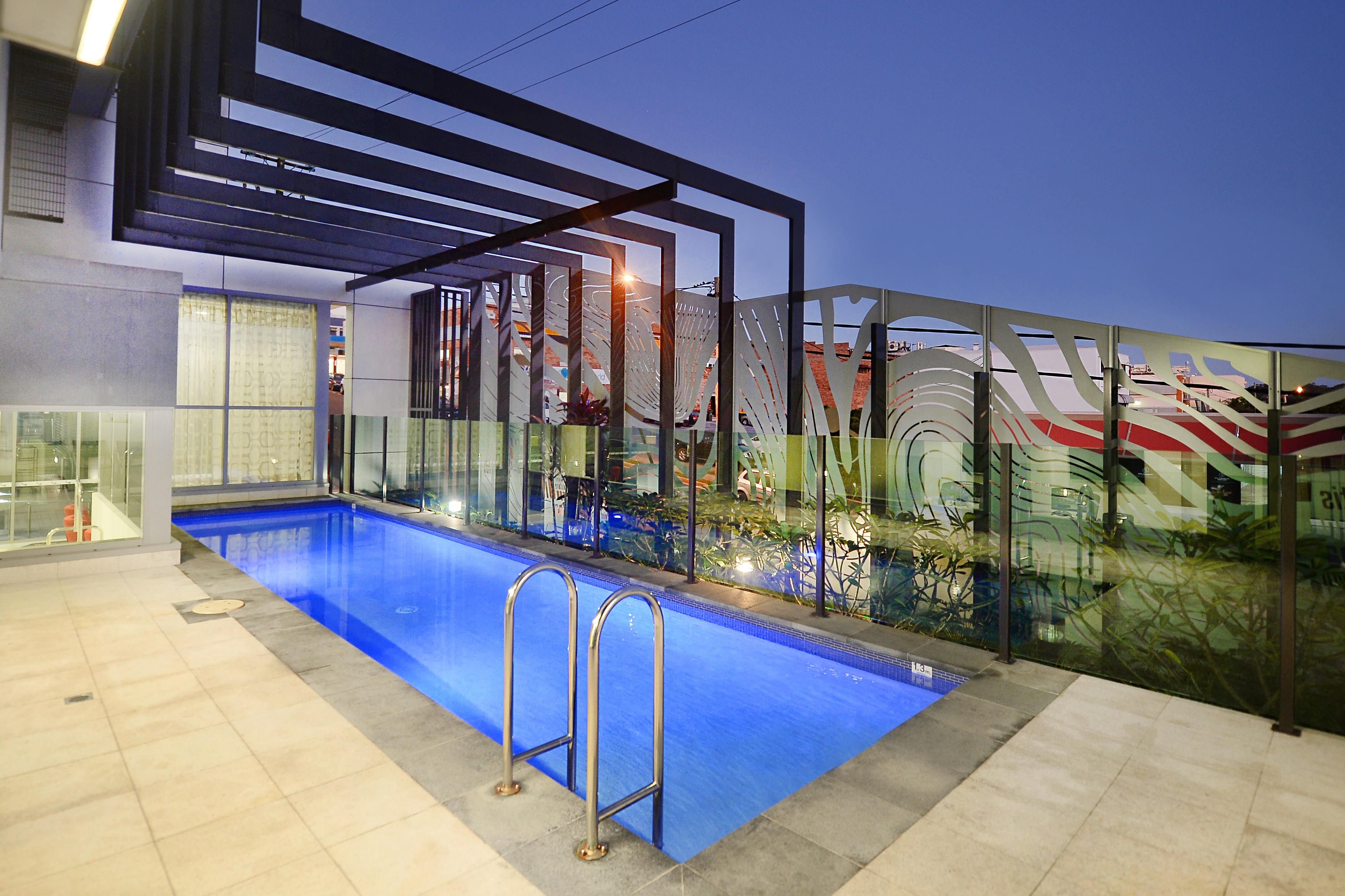 An outdoor swimming pool with a glass barrier, metal railings, and a view of buildings in the background.