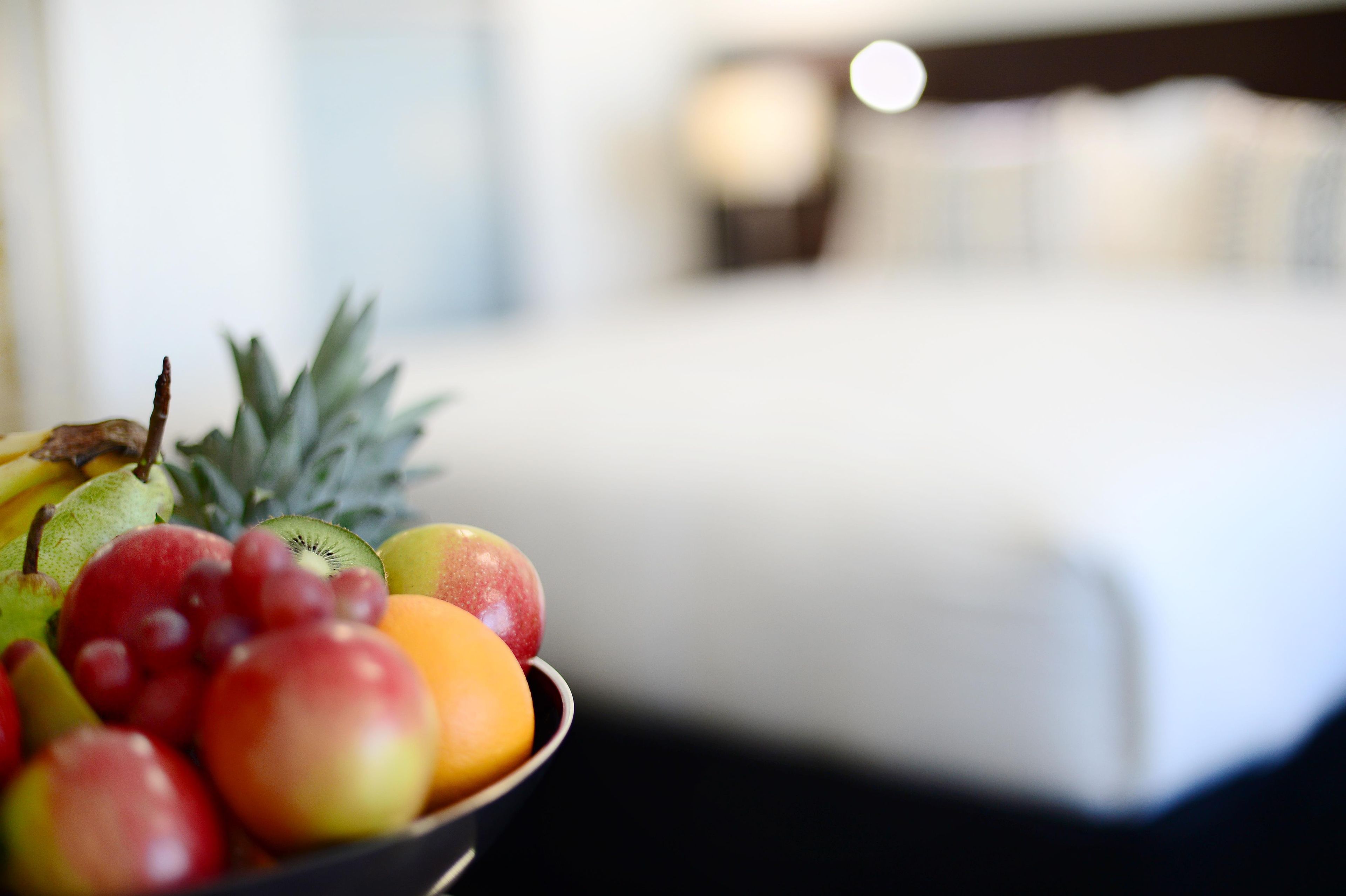 A fruit bowl on a table with a blurred background of a bed and a nightstand.