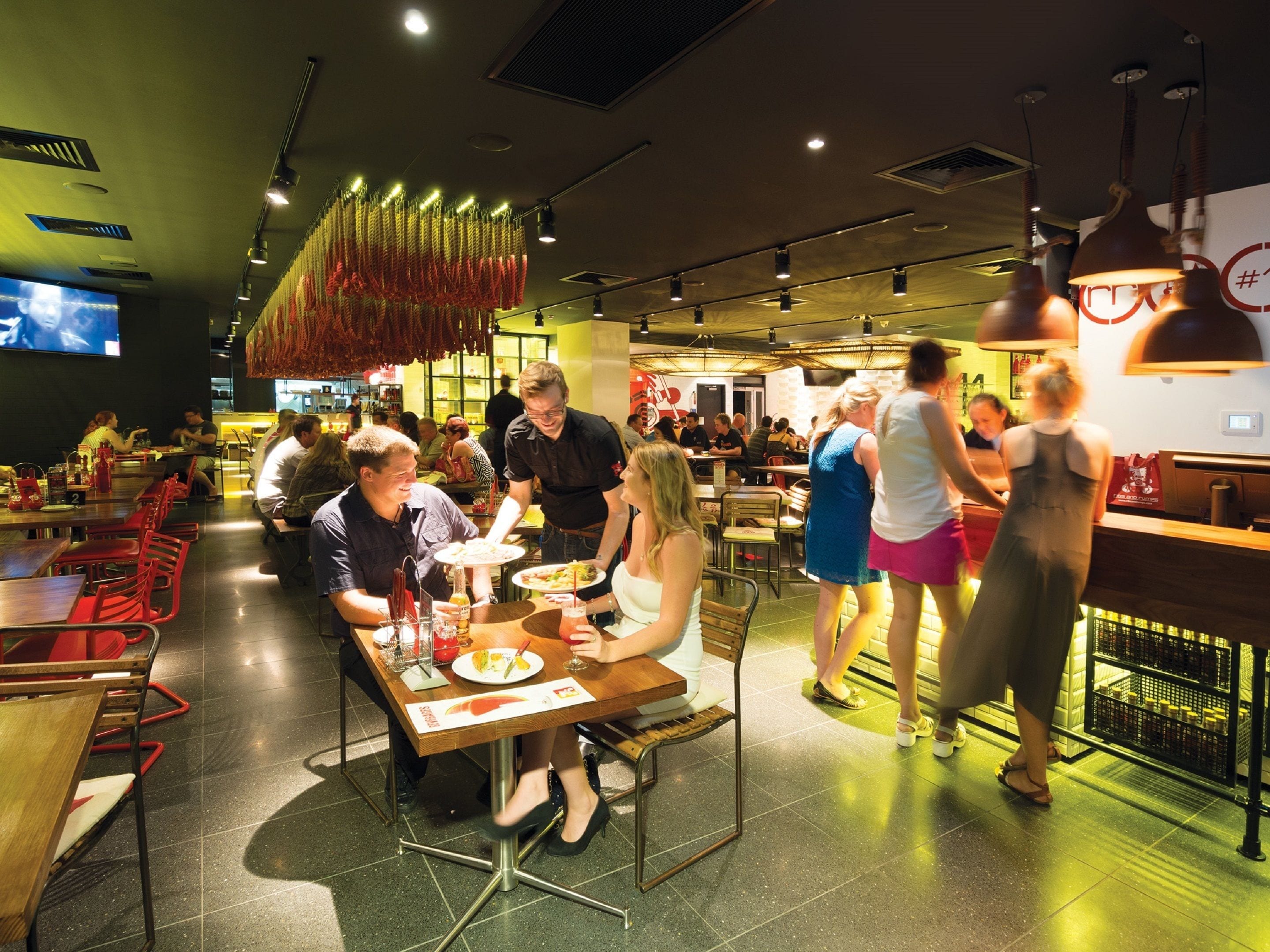 A restaurant with multiple dining tables and chairs, a bar area, and a television screen on the wall.