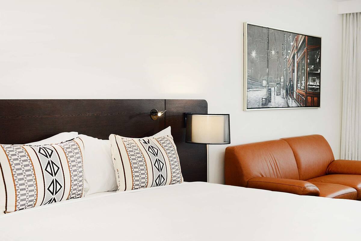 Een hotelkamer met een bed, een nachtkastje, een lamp, een bruine leren fauteuil en een stadsgezicht schilderij aan de muur.