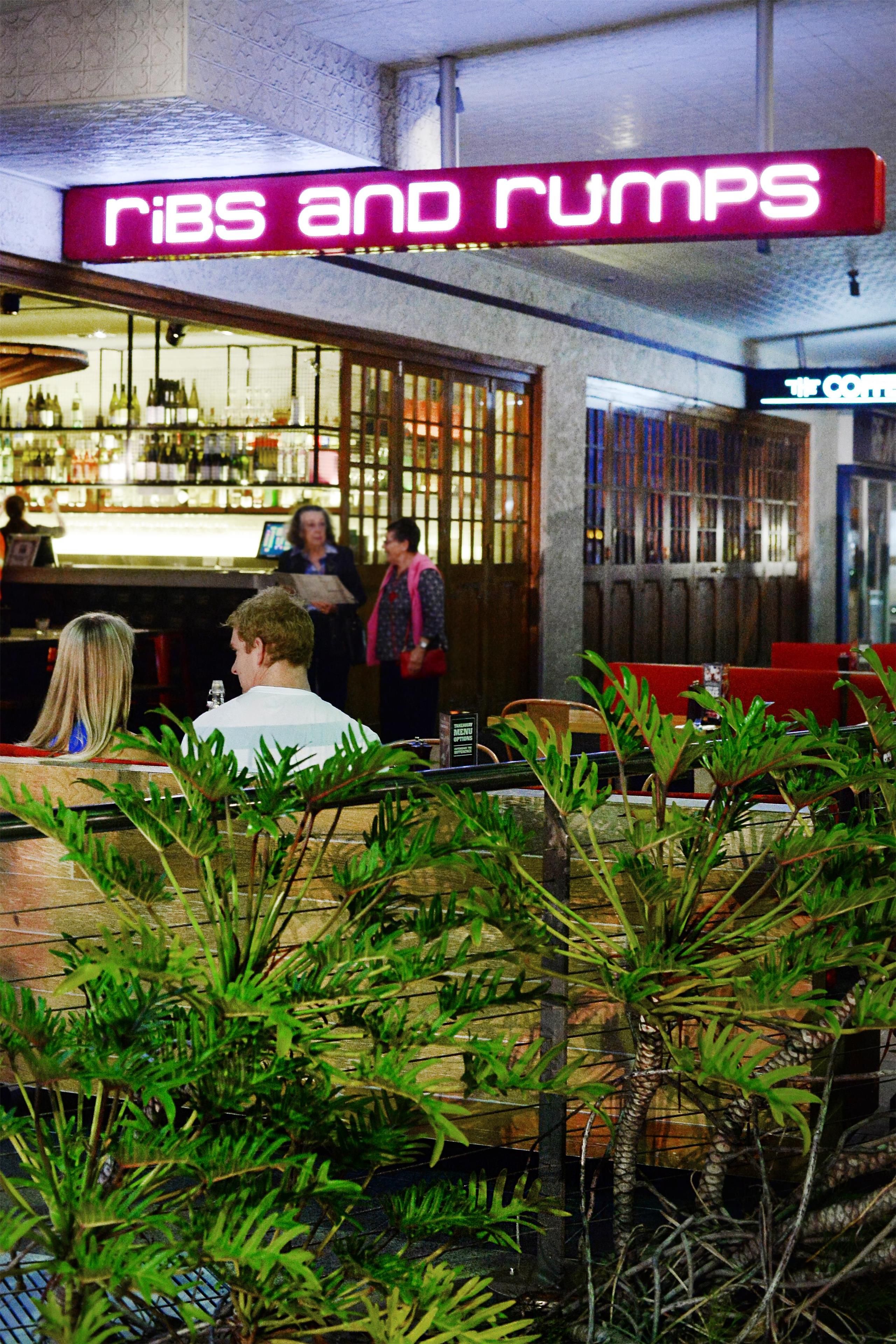 Zona de bar con un letrero de neón que dice 'Ribs and Rumps' y una pizarra de menú.