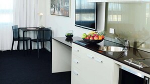 Una cocina moderna con encimera negra, armarios blancos, un fregadero de acero inoxidable y un bol con fruta. Hay una zona de comedor con una mesa y sillas, y un cuadro en la pared.
