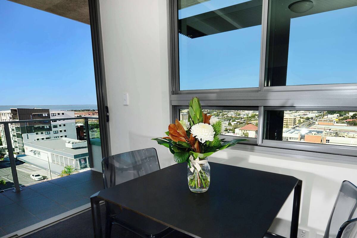 Una habitación moderna con un amplio ventanal que ofrece vistas a la ciudad, una mesa negra y un jarrón con flores.