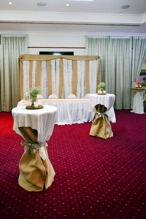 Een balzaal met twee ronde tafels, bedekt met witte tafelkleden, elk met een kaars en een kleine plant. De zaal heeft een rood tapijt en is versierd met een gestreepte gordijnen achtergrond.