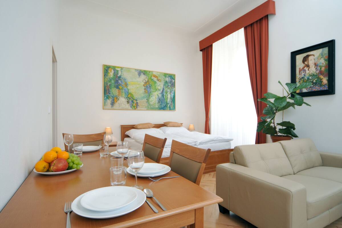 A hotel room with a dining table set for two, a bed with a headboard, a sofa, a potted plant, and two paintings on the walls.