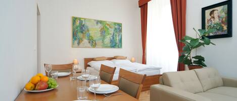 A hotel room with a dining table set for two, a bed with a headboard, a sofa, a potted plant, and two paintings on the walls.
