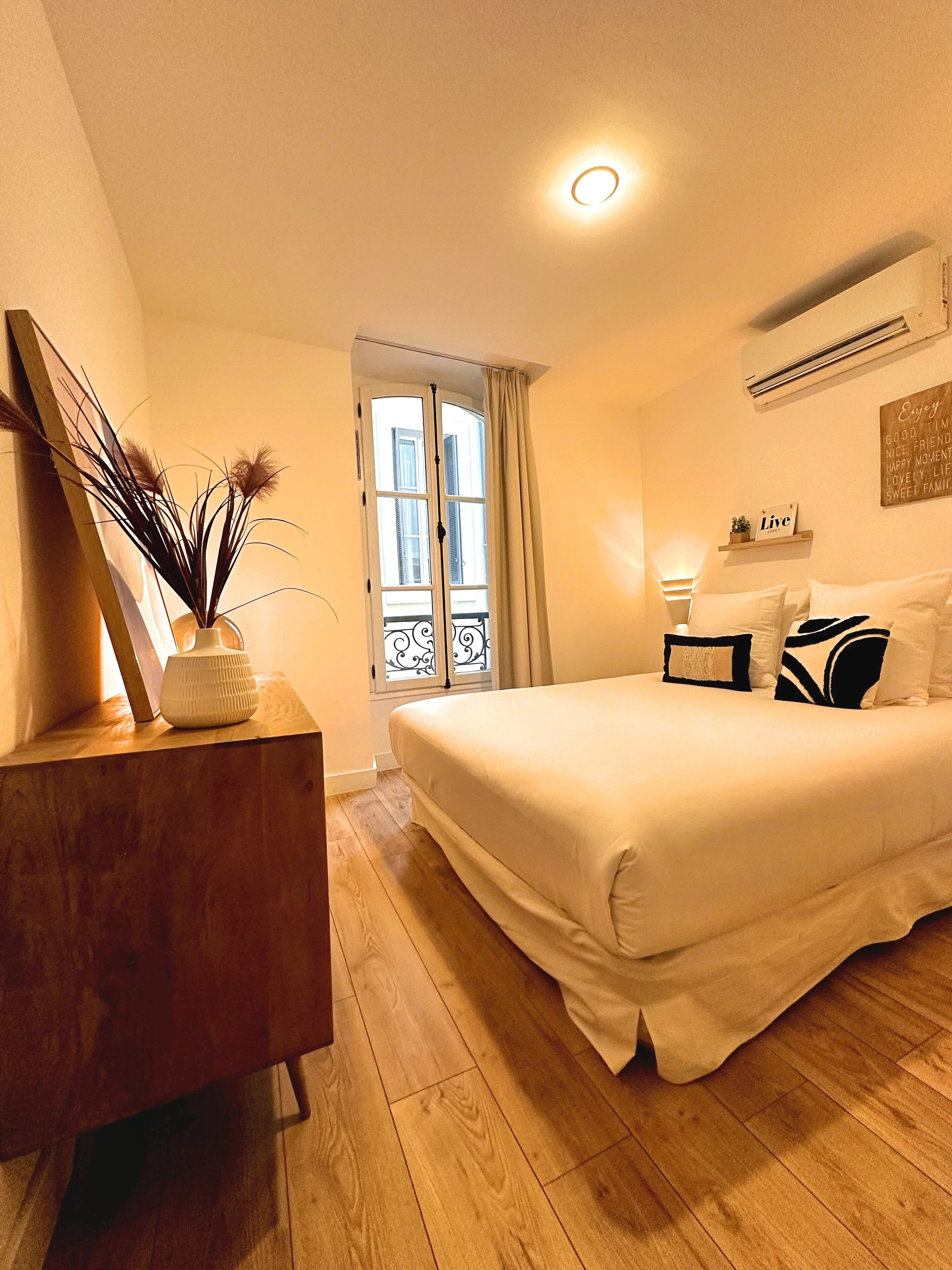 Un dormitorio con cama, cómoda, ventana con cortinas, aire acondicionado de pared y mesita de noche con lámpara.